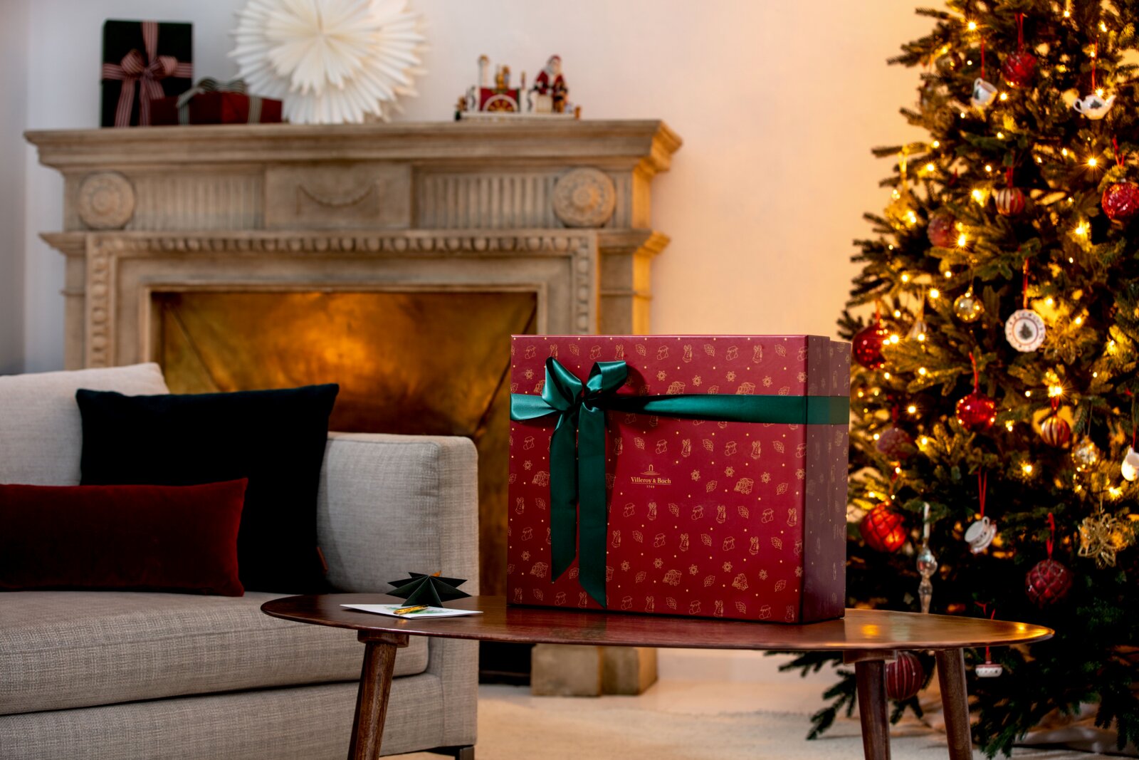 Große rote Villeroy & Boch Geschenkbox mit goldenen Weihnachtsmotiven und dunkelgrüner Schleife, frontal in leichter Seitenperspektive auf einem Couchtisch vor Kamin und festlich beleuchtetem Weihnachtsbaum präsentiert