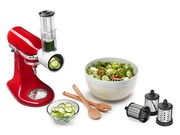 Roter Gemüseschneider mit Schnitzwerk, von der Seite gezeigt, neben einer Schüssel mit frischem Salat und verschiedenen Schneideaufsätzen.