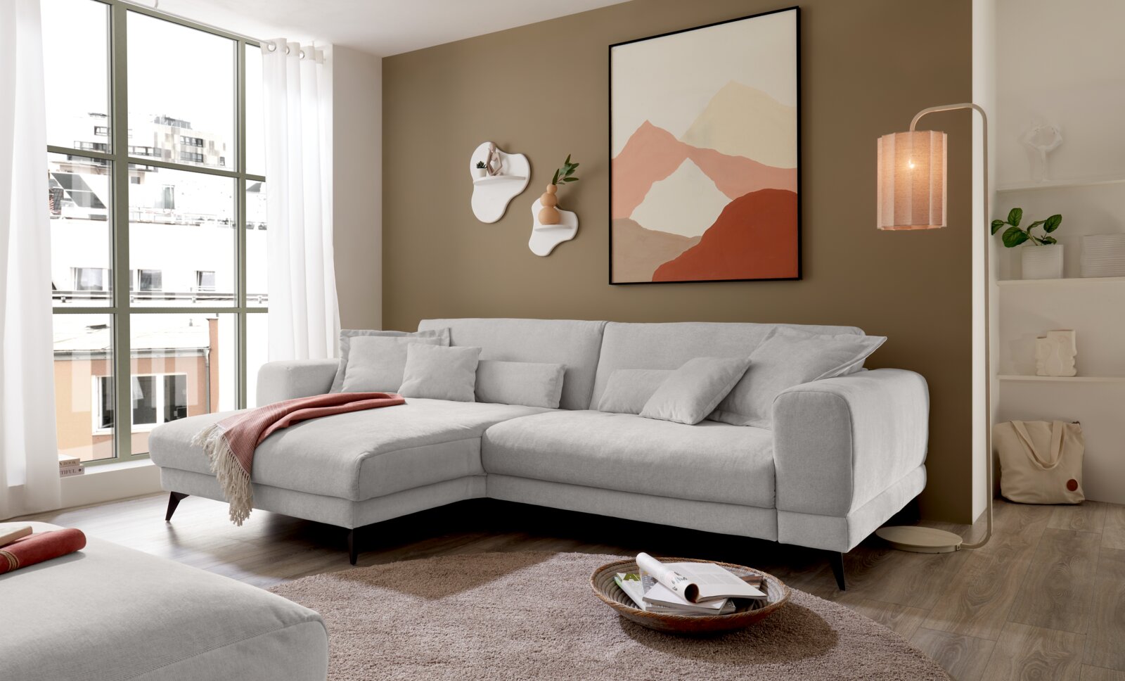 Set one by Musterring Ecksofa SET ONE MIDLAND Modernes hellgraues L-förmiges Ecksofa mit Recamiere links und mehreren Kissen in einem hellen Wohnzimmer, fotografiert aus schräger Frontperspektive.