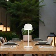 Weiße, moderne LED-Tischleuchte mit rundem Schirm auf einem gedeckten Holztisch im Außenbereich, frontal aus Augenhöhe aufgenommen.
