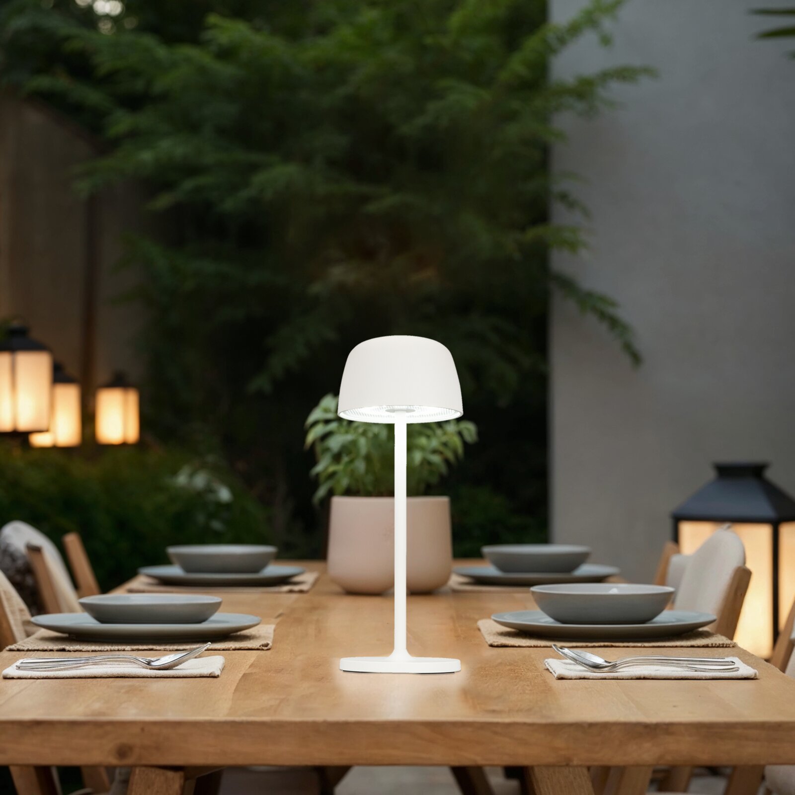 Weiße, moderne LED-Tischleuchte mit rundem Schirm auf einem gedeckten Holztisch im Außenbereich, frontal aus Augenhöhe aufgenommen.