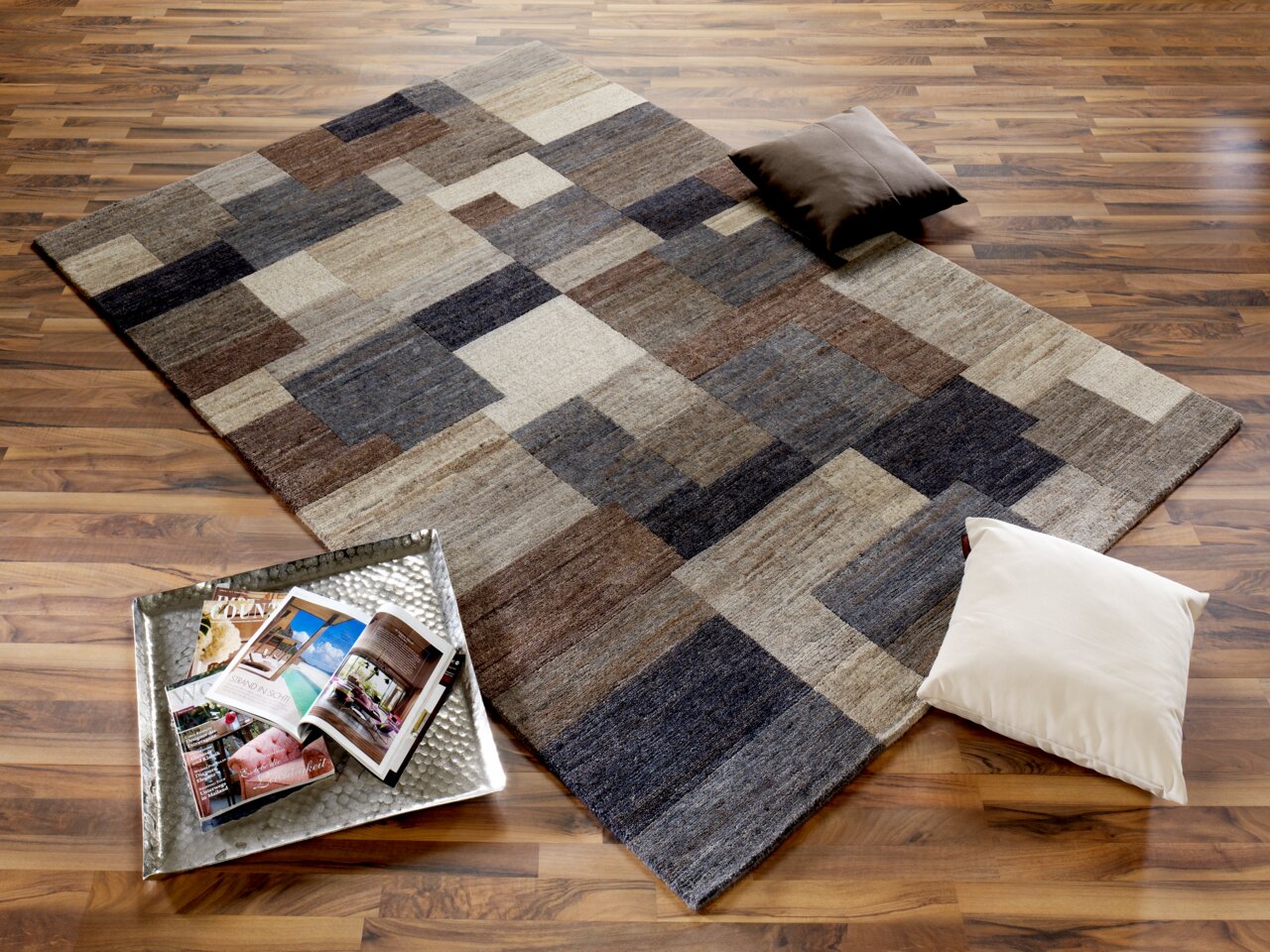Moderner Wollteppich mit geometrischem Muster in Erdtönen, auf einem Holzboden liegend, mit zwei Kissen und einem Tablett mit Zeitschriften. Perspektive von oben.