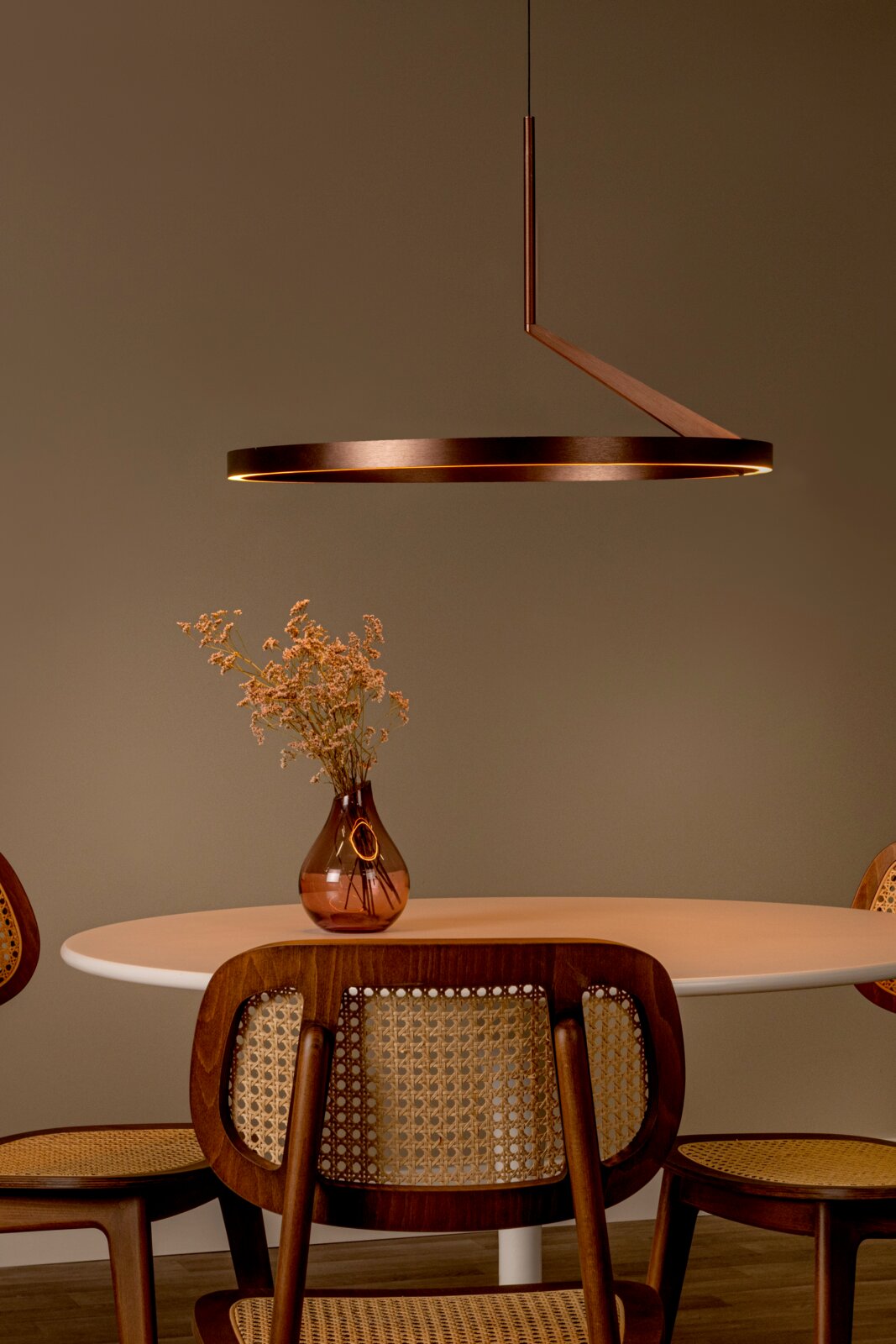 Moderne, ringförmige LED-Pendelleuchte in Bronze/Metalloptik über einem runden Esstisch, seitliche Perspektive mit leichtem Blick von vorn, warmes Licht in minimalistischer Wohnszene