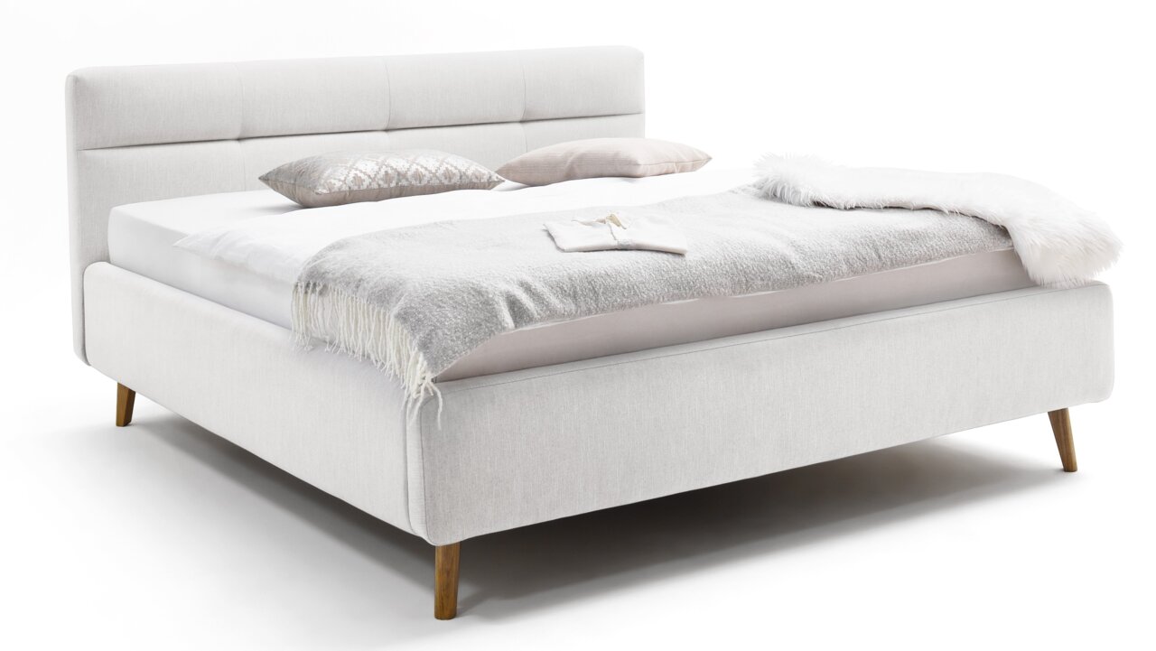Zijaanzicht van een elegant gestoffeerd bed in een lichte stof, met hoofdeinde en houten poten, versierd met kussens en een deken.
