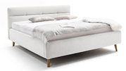 Zijaanzicht van een elegant gestoffeerd bed in een lichte stof, met hoofdeinde en houten poten, versierd met kussens en een deken.