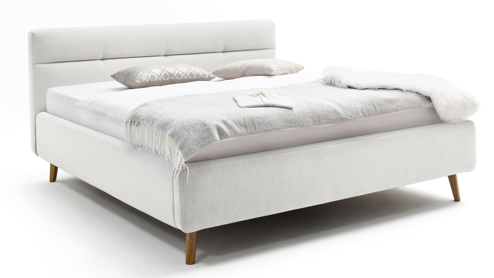 Seitliche Perspektive auf ein elegantes Polsterbett in hellem Stoff mit Kopfteil und Holzfüßen, dekoriert mit Kissen und einer Decke.