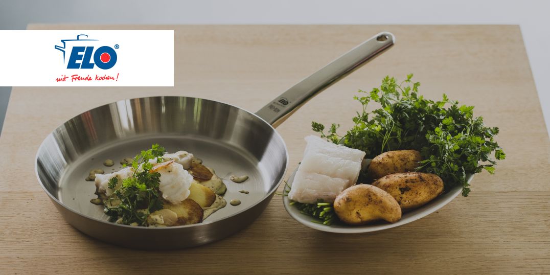 Een elegante roestvrijstalen pan met vis, kruiden en aardappelen, en verse ingrediënten op een wit bord.
