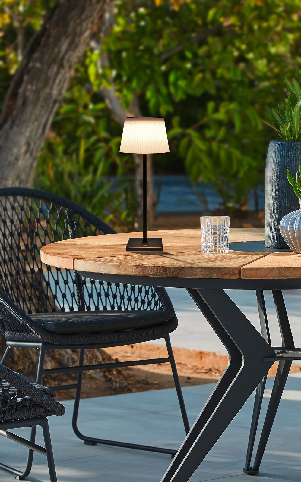 Ambia Garden Solarleuchte Solarleuchte auf einem Holztisch im Freien, umgeben von Gartenmöbeln und Pflanzen, seitliche Perspektive