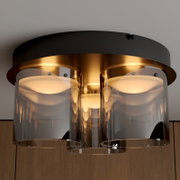 Moderne Deckenleuchte mit schwarzem Baldachin, goldfarbenem Innenkörper und drei rauchgrauen Glaszylindern, leuchtend in warmer Lichtfarbe, Nahaufnahme von unten (Untersicht)