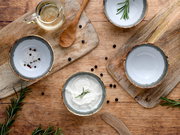 Draufsicht auf rustikales Food-Arrangement: Keramikschalen mit Salz und Pfefferkörnern sowie cremigem Dip mit Rosmarin, dazu Holzlöffel und Glas Öl auf Holzbrettchen und Holztisch.