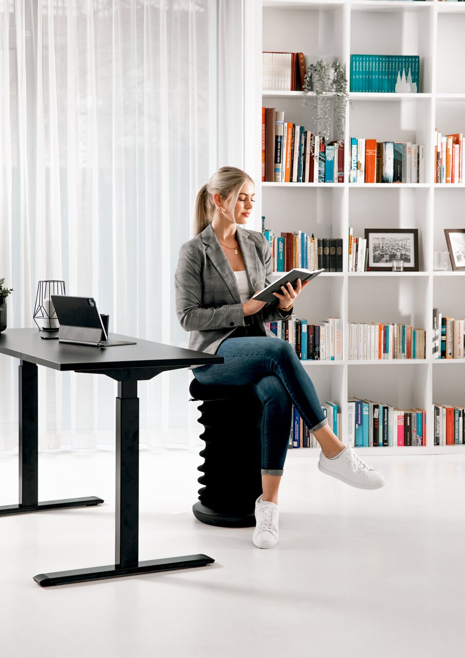 Moderner E-Schreibtisch in einem stilvollen Büro mit einer Frau, die auf einem ergonomischen Hocker sitzt und ein Buch liest. Seitliche Perspektive auf den Schreibtisch.
