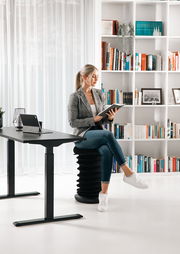 Moderner E-Schreibtisch in einem stilvollen Büro mit einer Frau, die auf einem ergonomischen Hocker sitzt und ein Buch liest. Seitliche Perspektive auf den Schreibtisch.