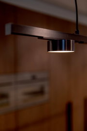 Close-up van een moderne hanglamp met metalen arm en cilindervormige spot die warm wit licht naar beneden schijnt, gefotografeerd vanuit een licht schuine zijaanzicht/perspectief.