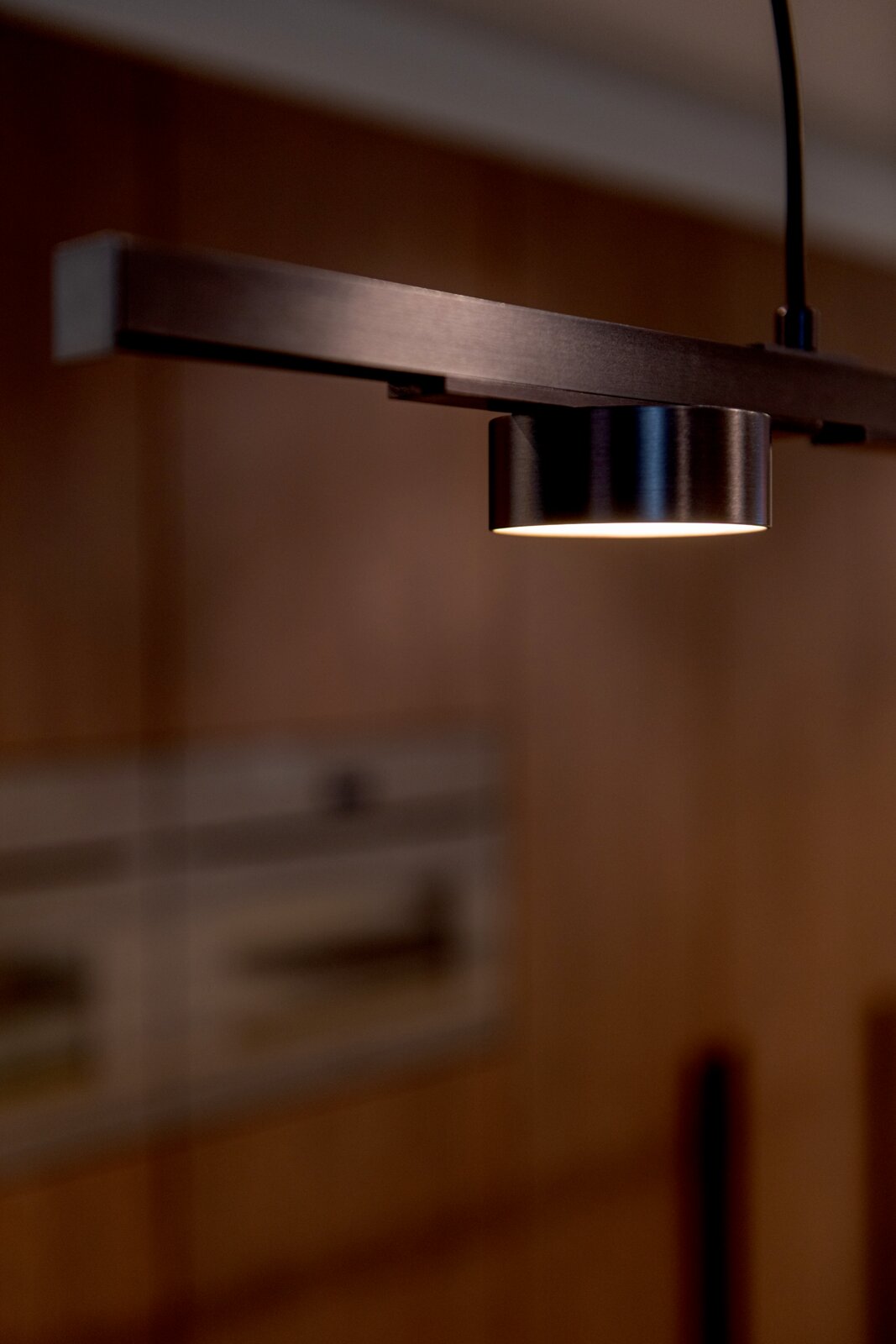 Close-up van een moderne hanglamp met metalen arm en cilindervormige spot die warm wit licht naar beneden schijnt, gefotografeerd vanuit een licht schuine zijaanzicht/perspectief.