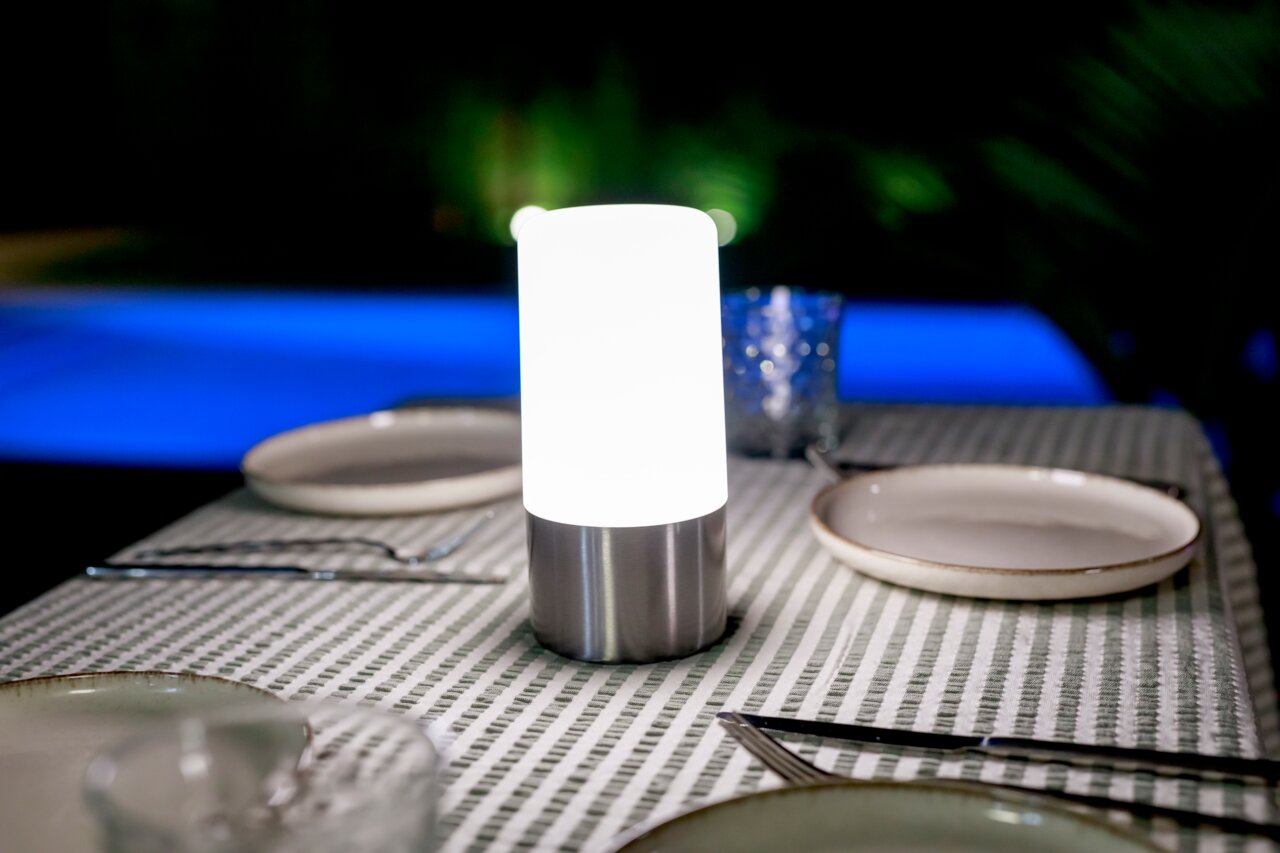 Newgarden Akku-Tischleuchte MAIA Leuchtende kabellose LED-Tischlampe mit weißem Diffusor und Edelstahlfuß auf gedecktem Esstisch, frontal aus Augenhöhe fotografiert.