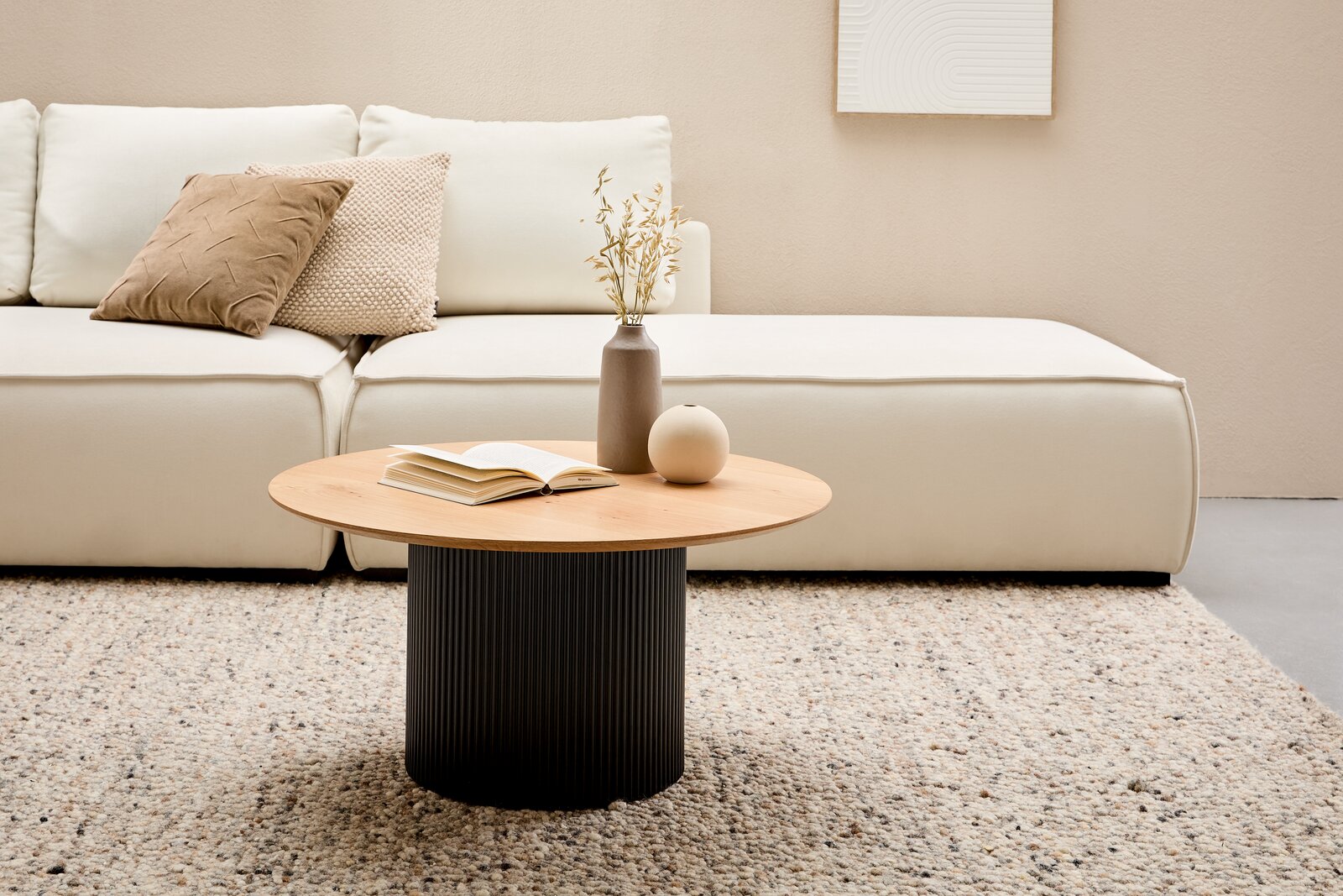 Moderner Couchtisch mit runder Holzplatte und geriffeltem schwarzen Sockel, im Wohnzimmer vor einem cremefarbenen Sofa, aus der Frontalperspektive.