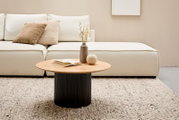 Moderner Couchtisch mit runder Holzplatte und geriffeltem schwarzen Sockel, im Wohnzimmer vor einem cremefarbenen Sofa, aus der Frontalperspektive.