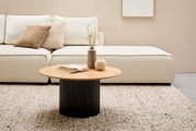 Moderne salontafel met een rond houten blad en een geribbelde zwarte voet, in de woonkamer voor een crèmekleurige bank, gezien van voren.
