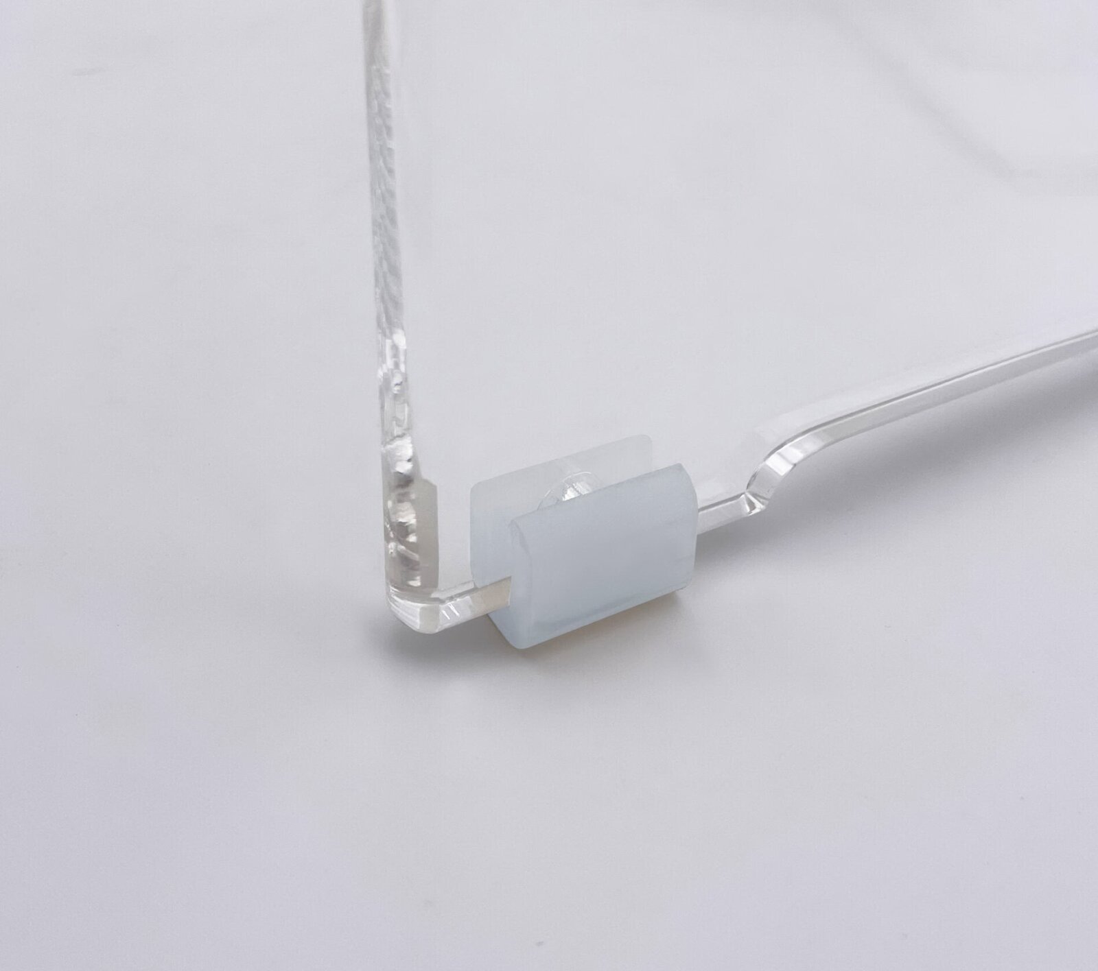 Nahaufnahme aus seitlicher Perspektive auf eine transparente Acrylglas-Platte mit milchig-weißem, selbstklebendem Silikon-Puffer/Abstandshalter an der unteren Ecke vor hellem Hintergrund