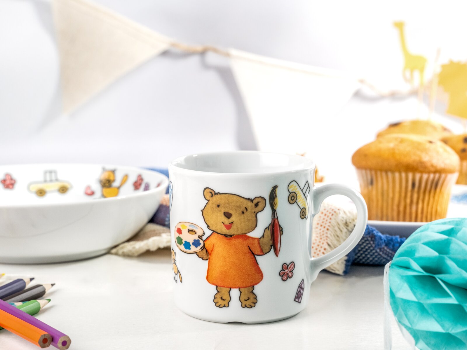 Weiße Kinder-Tasse aus Keramik mit Bären-Illustration (Bär im orangefarbenen Shirt mit Malpalette und Pinsel), frontal aus leicht seitlicher Perspektive fotografiert; im Hintergrund Schale und Muffins auf hellem Tisch.