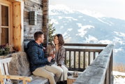 Ein Paar sitzt auf einer Holzbank auf einer Terrasse mit Blick auf schneebedeckte Berge und hält Sektkelche in der Hand.