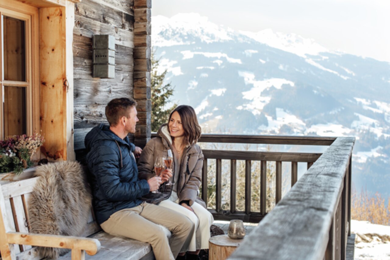 Ein Paar sitzt auf einer Holzbank auf einer Terrasse mit Blick auf schneebedeckte Berge und hält Sektkelche in der Hand.