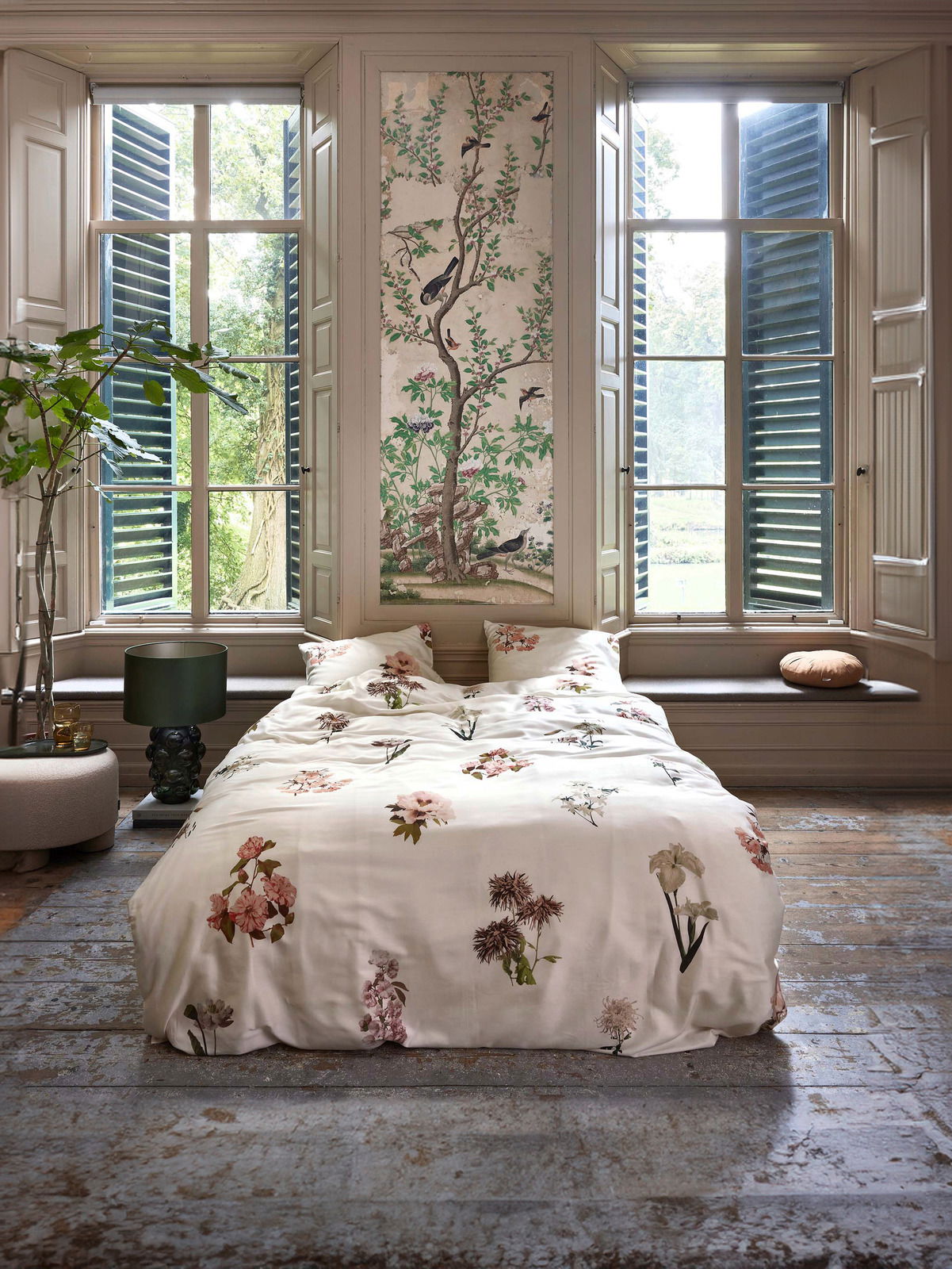 Bettwäsche mit floralen Mustern in einem stilvollen Raum mit großen Fenstern und grüner Aussicht, aus der Perspektive vom Fußende des Bettes.