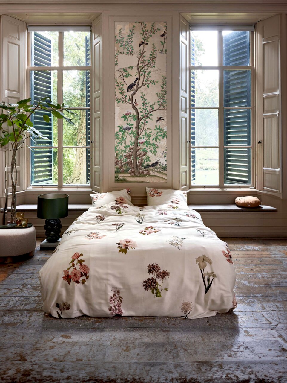 Bettwäsche mit floralen Mustern in einem stilvollen Raum mit großen Fenstern und grüner Aussicht, aus der Perspektive vom Fußende des Bettes.