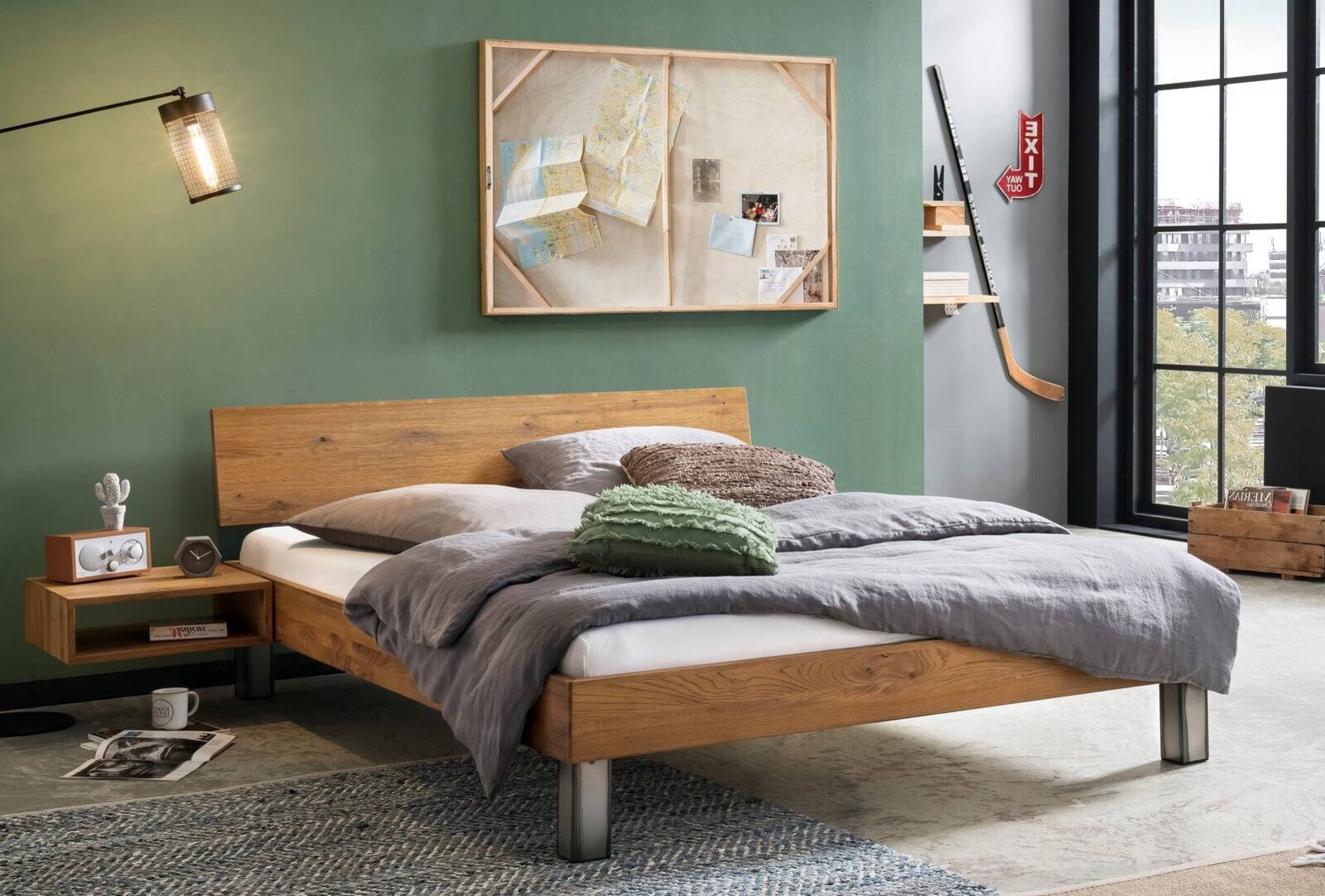 Modernes Doppelbett aus massivem Holz mit niedrigem Rahmen und Metallfüßen, im Industrial-Schlafzimmer mit grüner Wand und großer Fensterfront, perspektivisch schräg von vorne rechts aufgenommen