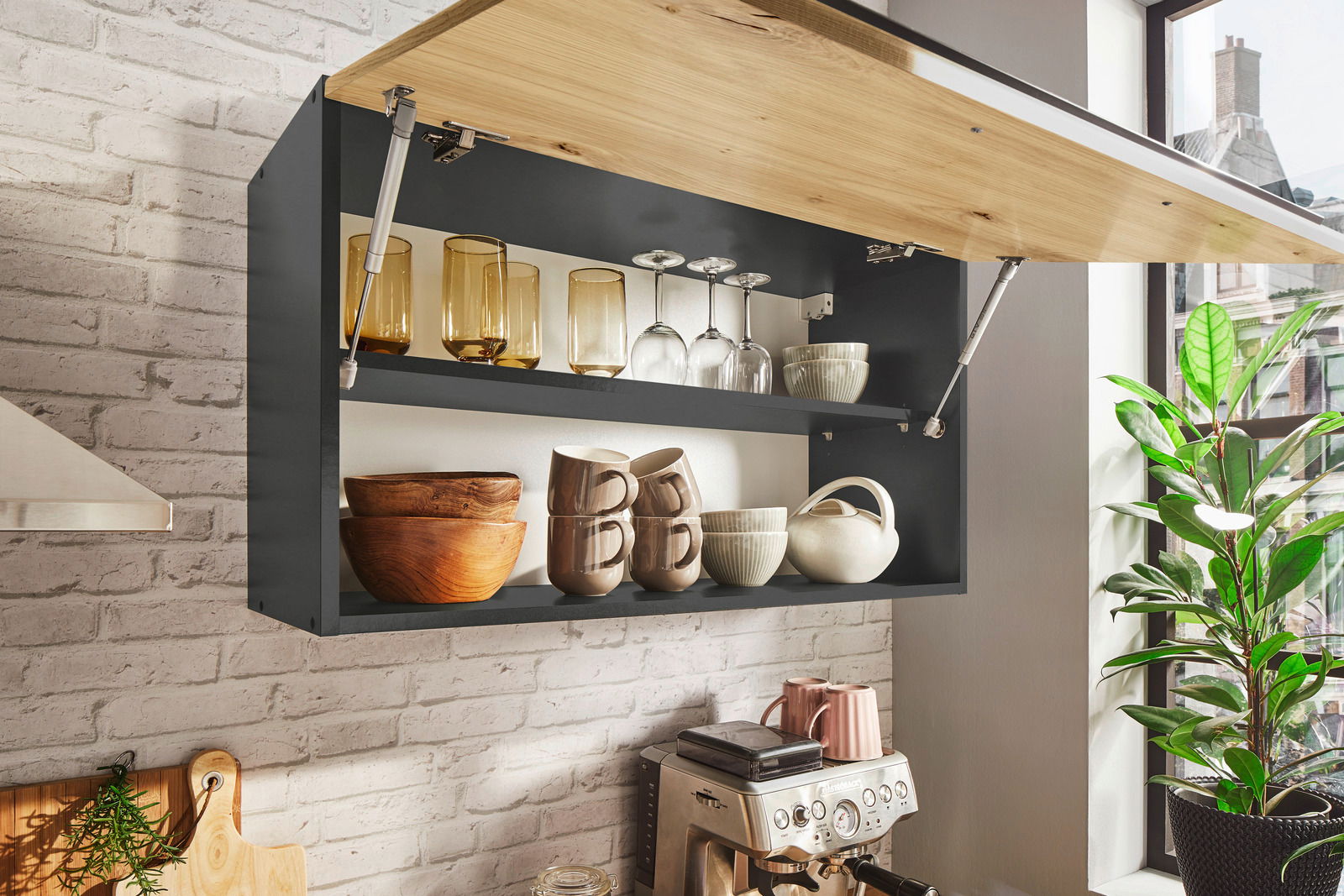 Küchenblock mit geöffnetem Hängeschrank, gefüllt mit Gläsern, Tassen und Schalen, in einer modernen Küche mit Backsteinwand und Espressomaschine, seitliche Perspektive.