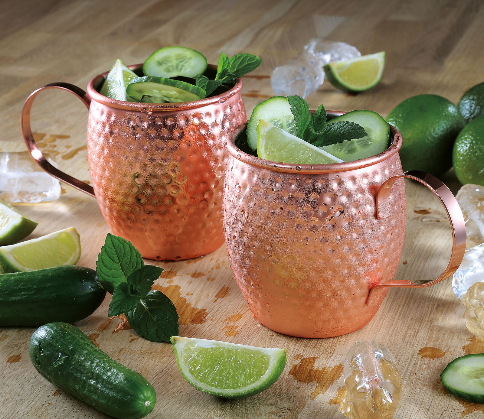 Zwei kupferfarbene Moscow Mule Becher mit gehämmertem Design, gefüllt mit Eis, Gurkenscheiben, Limetten und Minzblättern, auf einem Holztisch mit Limettenstücken und Gurken, aus seitlicher Perspektive.
