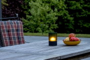 Akku-Windlicht auf einem Holztisch im Freien, neben einer Schale mit Äpfeln, aus seitlicher Perspektive fotografiert.