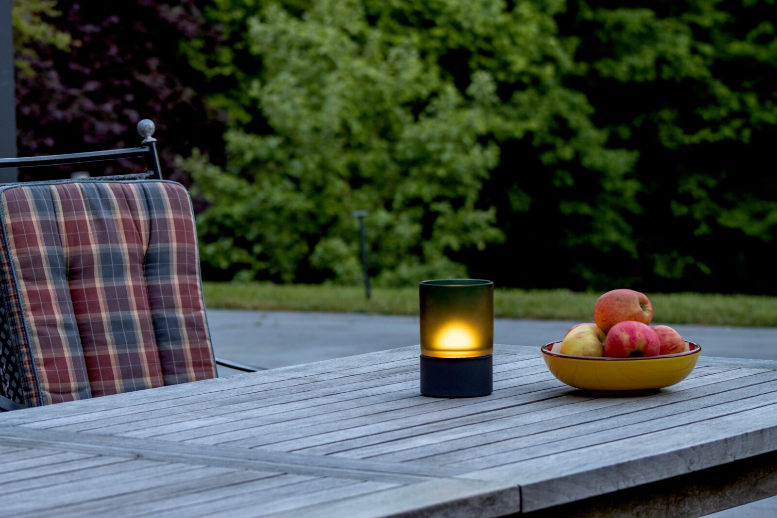 Akku-Windlicht auf einem Holztisch im Freien, neben einer Schale mit Äpfeln, aus seitlicher Perspektive fotografiert.
