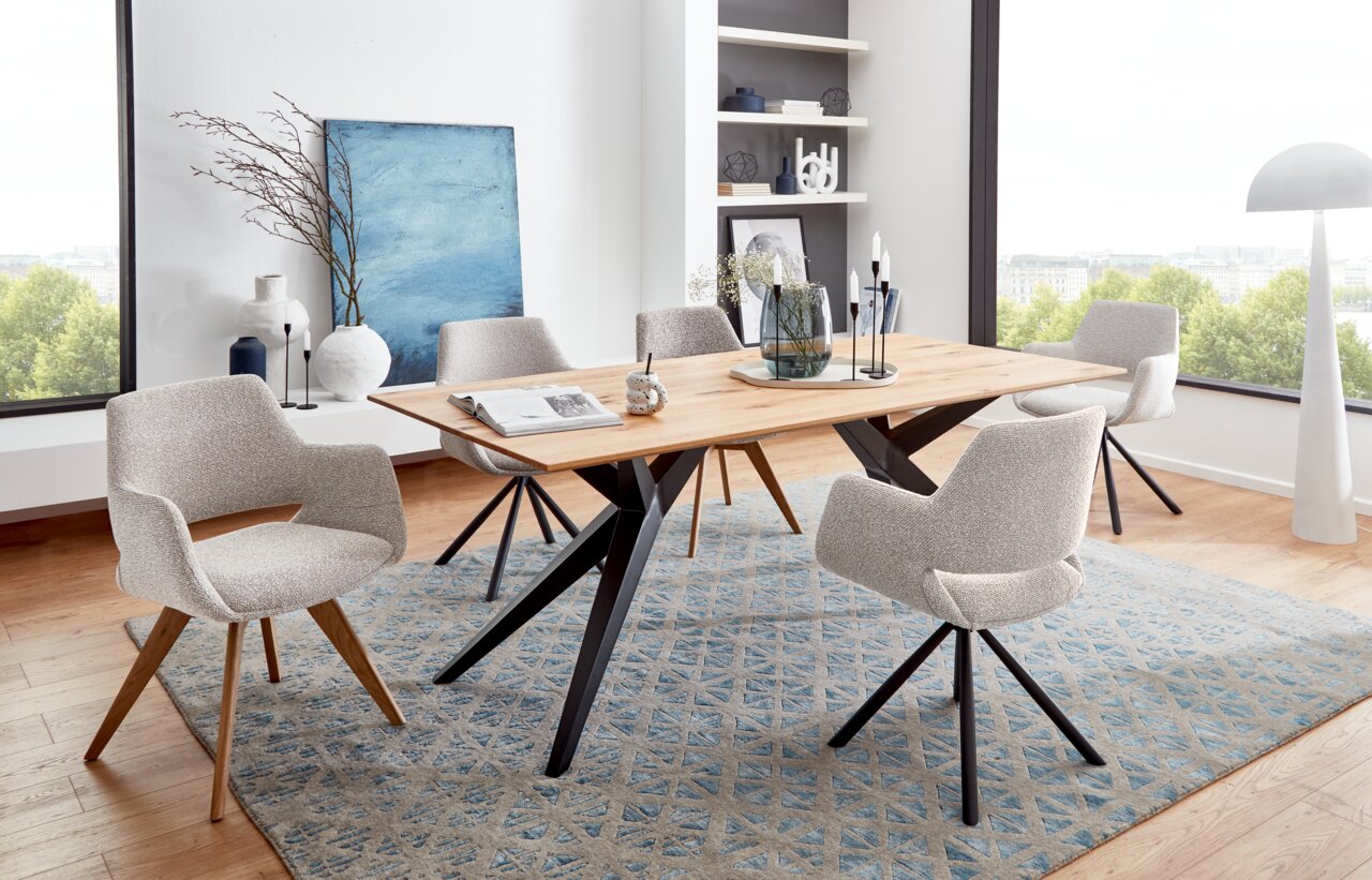 Musterring Fauteuil MR TARIO Moderne eettafel met houten blad en zwarte metalen poten, omringd door gestoffeerde stoelen met armleuningen in een lichte eetkamer met grote ramen. Perspectief: zijaanzicht.