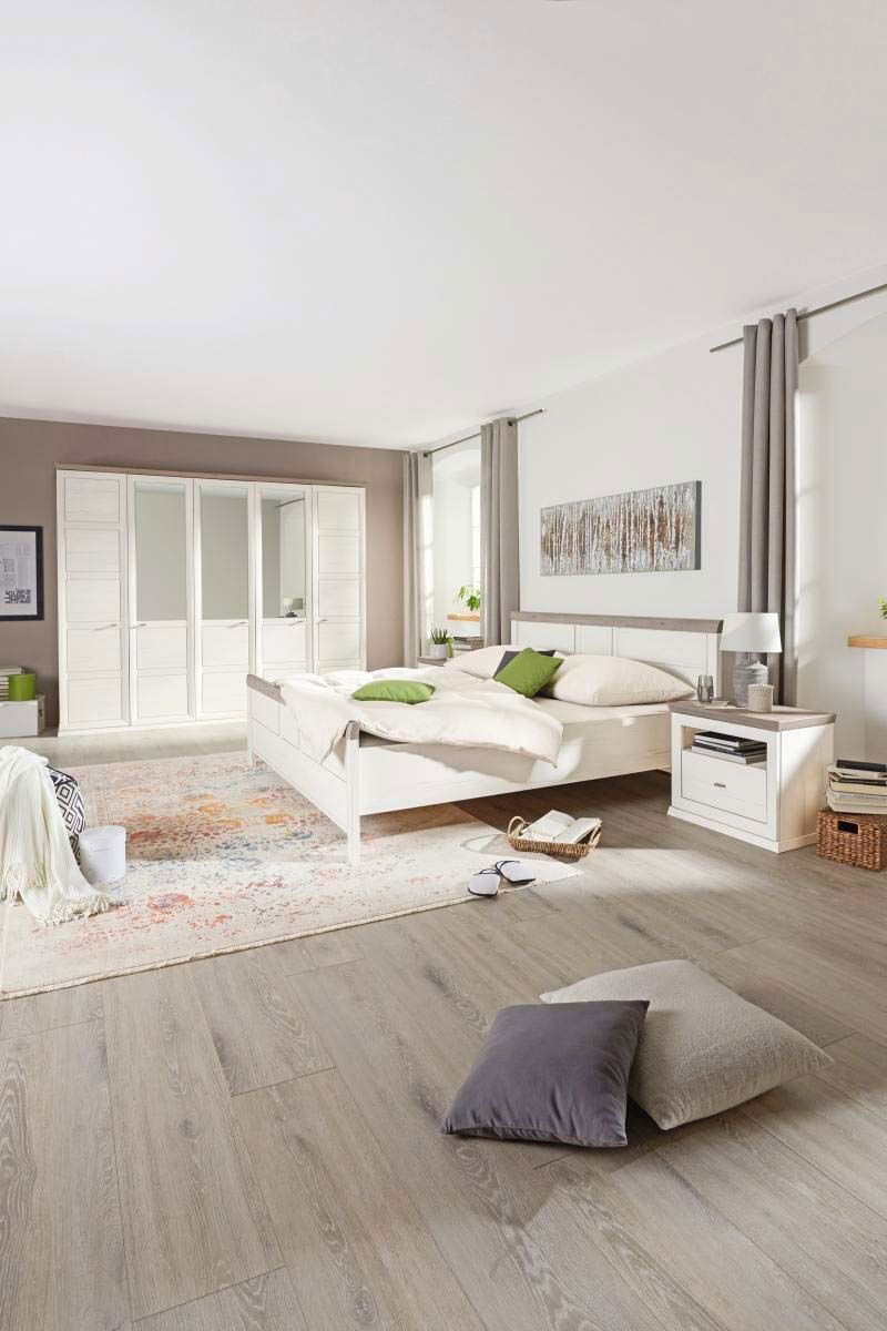 Schlafzimmer mit einem 5-türigen Drehtürenschrank CAMRON in heller Holzoptik, einem Doppelbett und einem Nachttisch, aus der Perspektive von vorne rechts betrachtet.