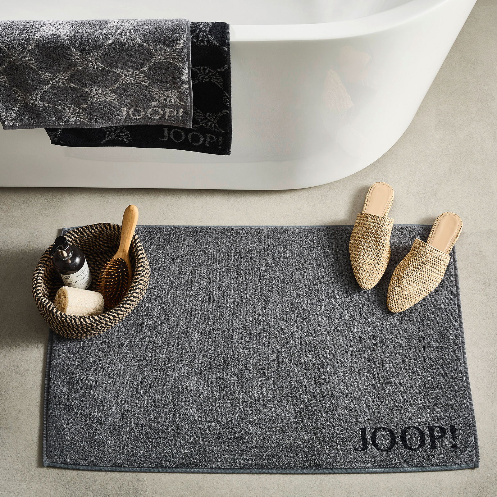 Graue Badematte mit 'JOOP!' Schriftzug, von oben fotografiert, neben einer Badewanne mit Handtüchern und einem Korb mit Badutensilien.