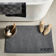 Graue Badematte mit 'JOOP!' Schriftzug, von oben fotografiert, neben einer Badewanne mit Handtüchern und einem Korb mit Badutensilien.