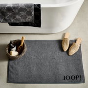 Graue Badematte mit 'JOOP!' Schriftzug, von oben fotografiert, neben einer Badewanne mit Handtüchern und einem Korb mit Badutensilien.