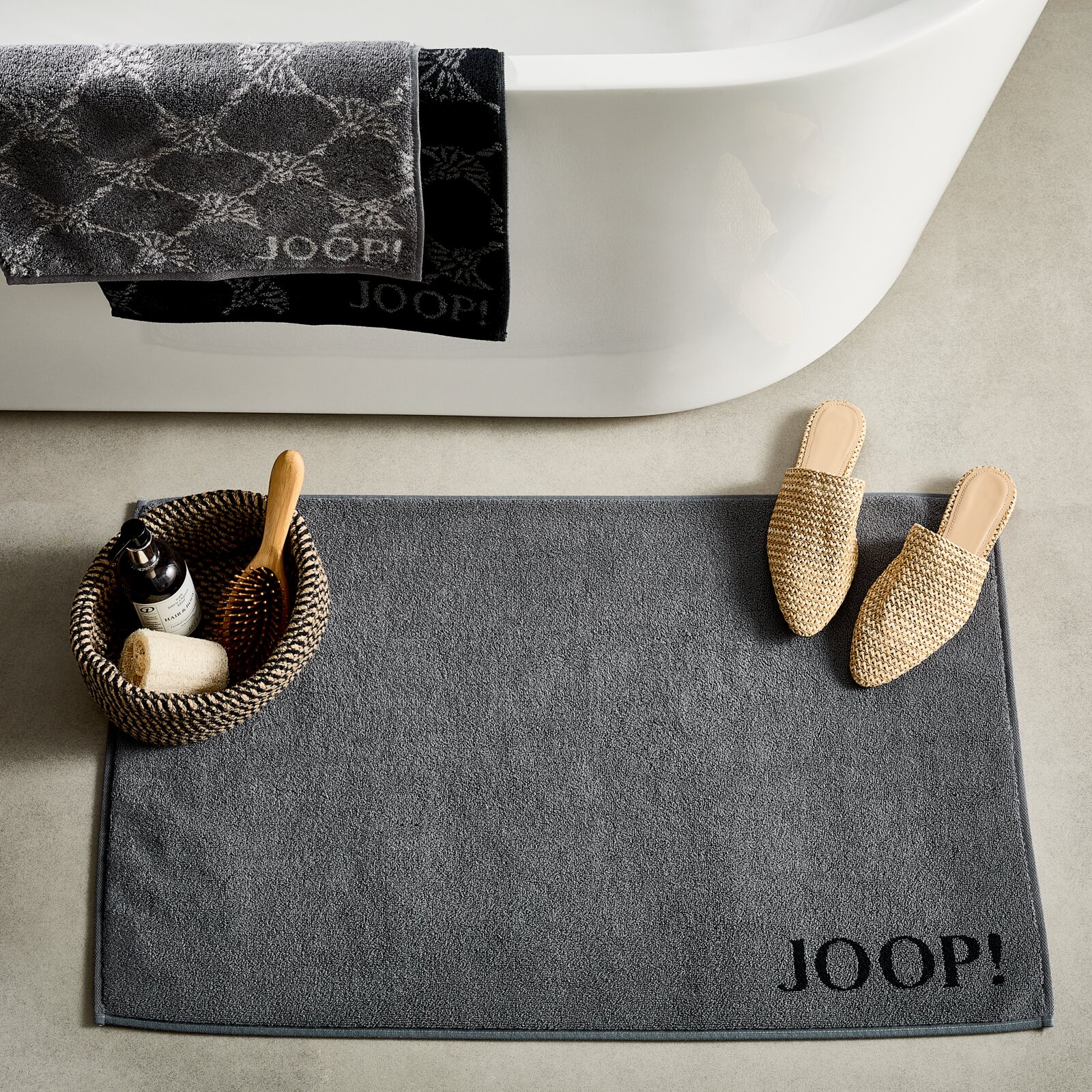 JOOP! Badematte DOUBLEFACE Graue Badematte mit 'JOOP!' Schriftzug, von oben fotografiert, neben einer Badewanne mit Handtüchern und einem Korb mit Badutensilien.