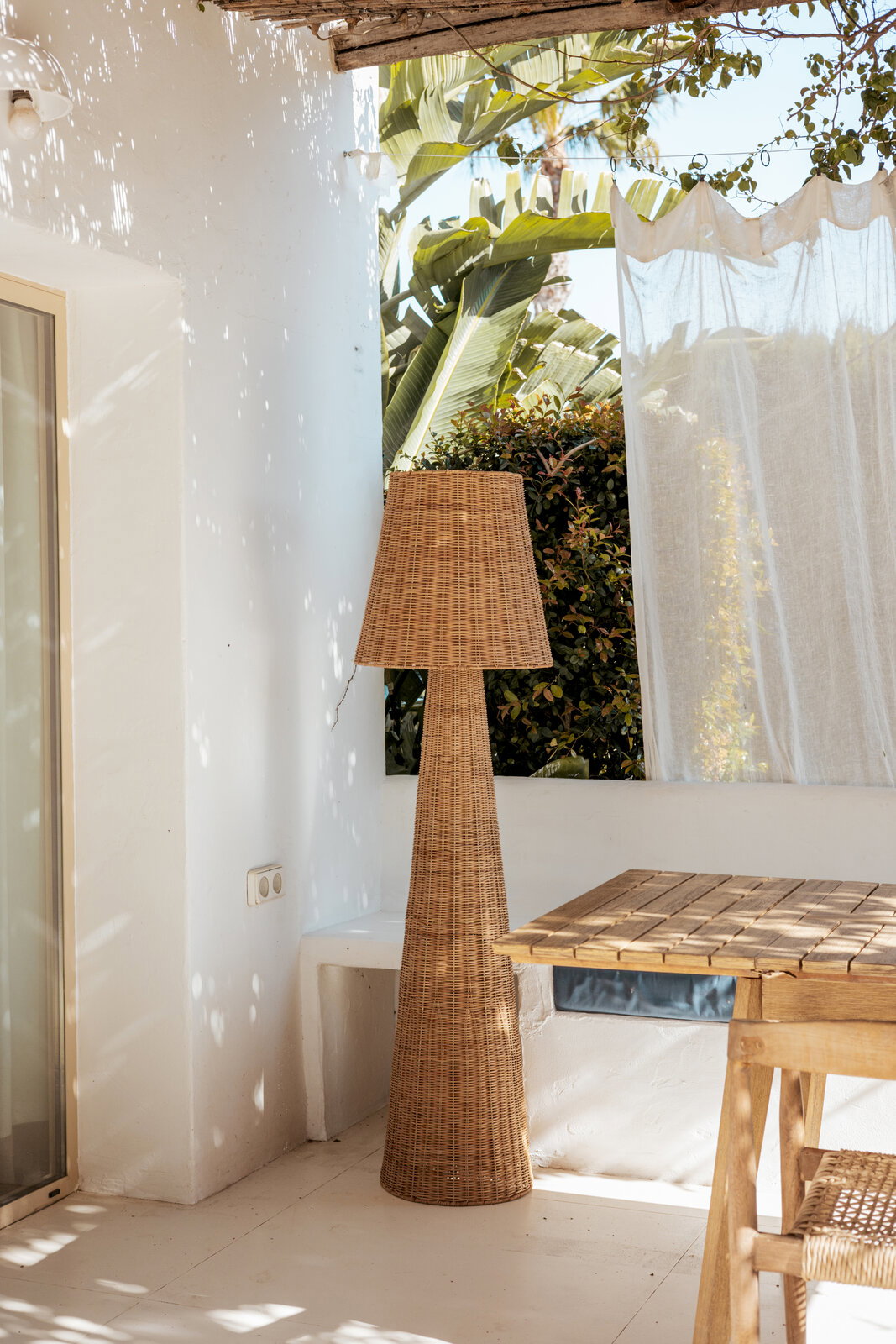 Hohe Stehlampe aus Rattan mit konischem Schirm auf einer hellen Terrasse neben Holztisch und weißer Wand, seitliche Perspektive auf das Produkt.
