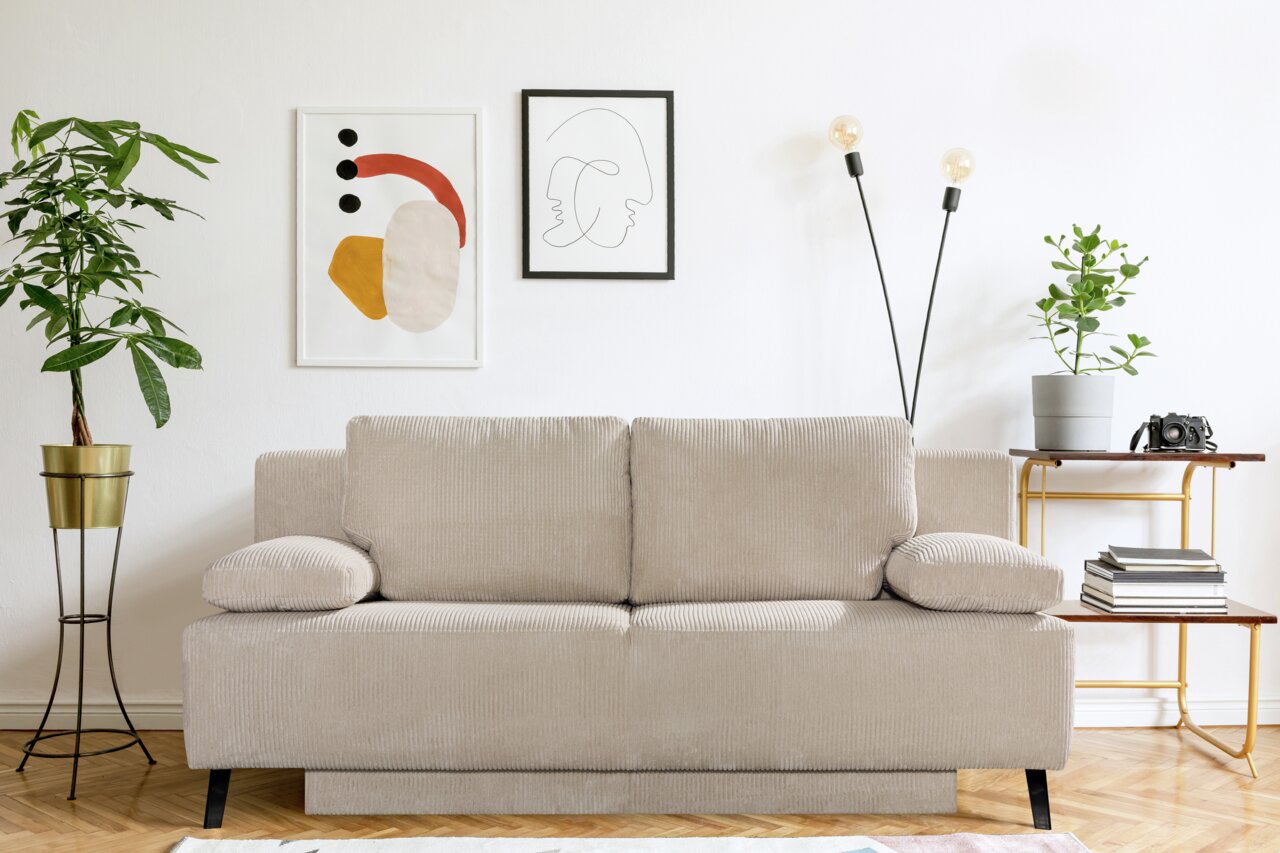 Beige Querschläfer-Sofa in einem modernen Wohnzimmer, frontal fotografiert. Umgeben von Pflanzen, Kunstwerken und einem Beistelltisch.