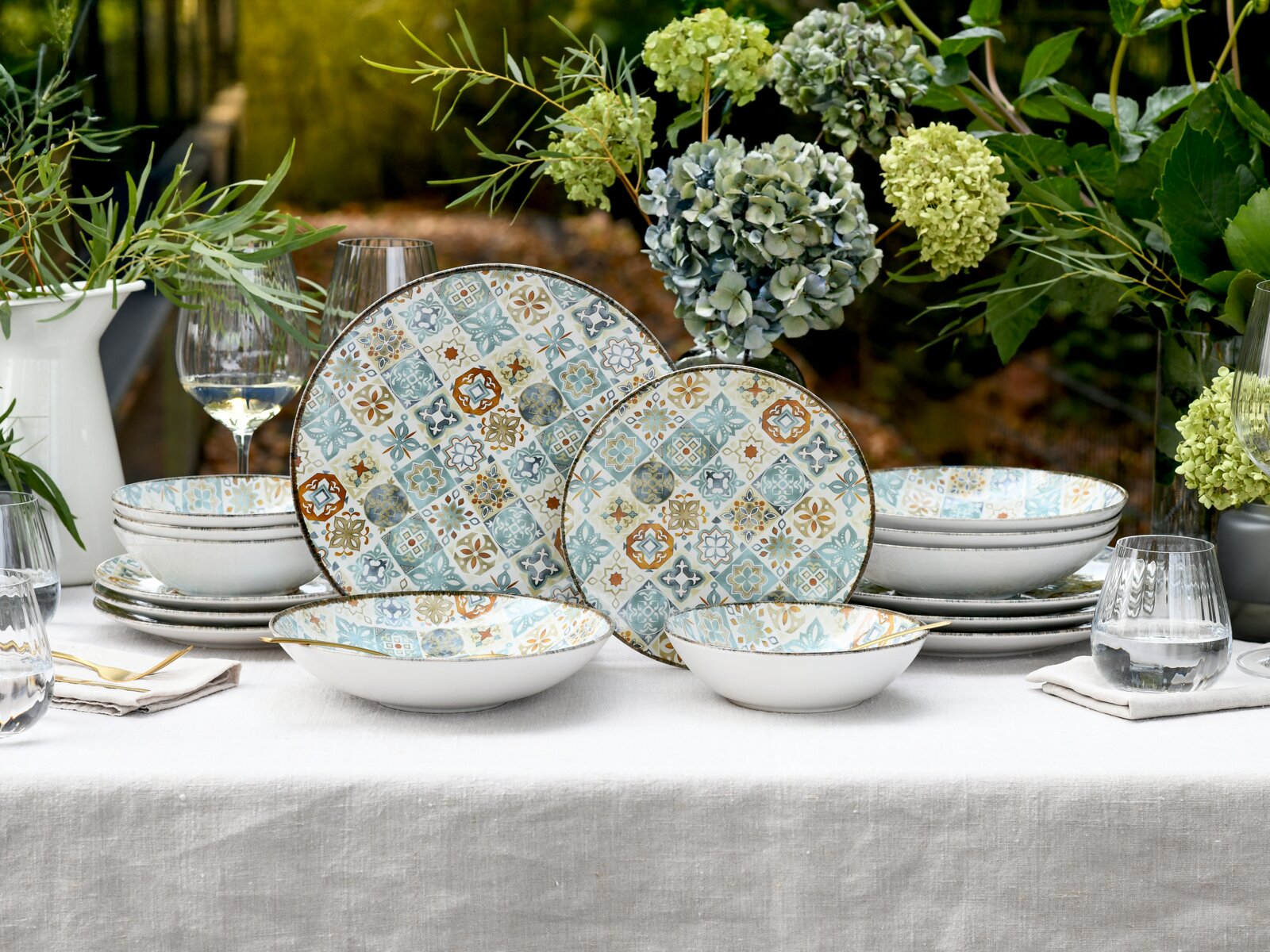 Frontale Tischszene: gemustertes Keramik-Geschirrset mit Tellern und Schalen im mediterranen Kachel-Design in Blau-, Beige- und Ockertönen, dekoriert auf weißer Tischdecke mit Gläsern und Hortensien im Garten