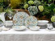 Frontale Tischszene: gemustertes Keramik-Geschirrset mit Tellern und Schalen im mediterranen Kachel-Design in Blau-, Beige- und Ockertönen, dekoriert auf weißer Tischdecke mit Gläsern und Hortensien im Garten