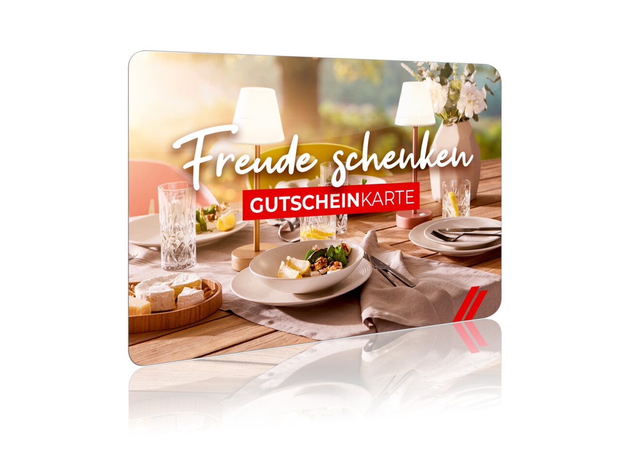 Gutscheinkarte mit der Aufschrift 'Freude schenken' auf einem gedeckten Tisch mit Tellern, Gläsern und Blumen, seitliche Perspektive