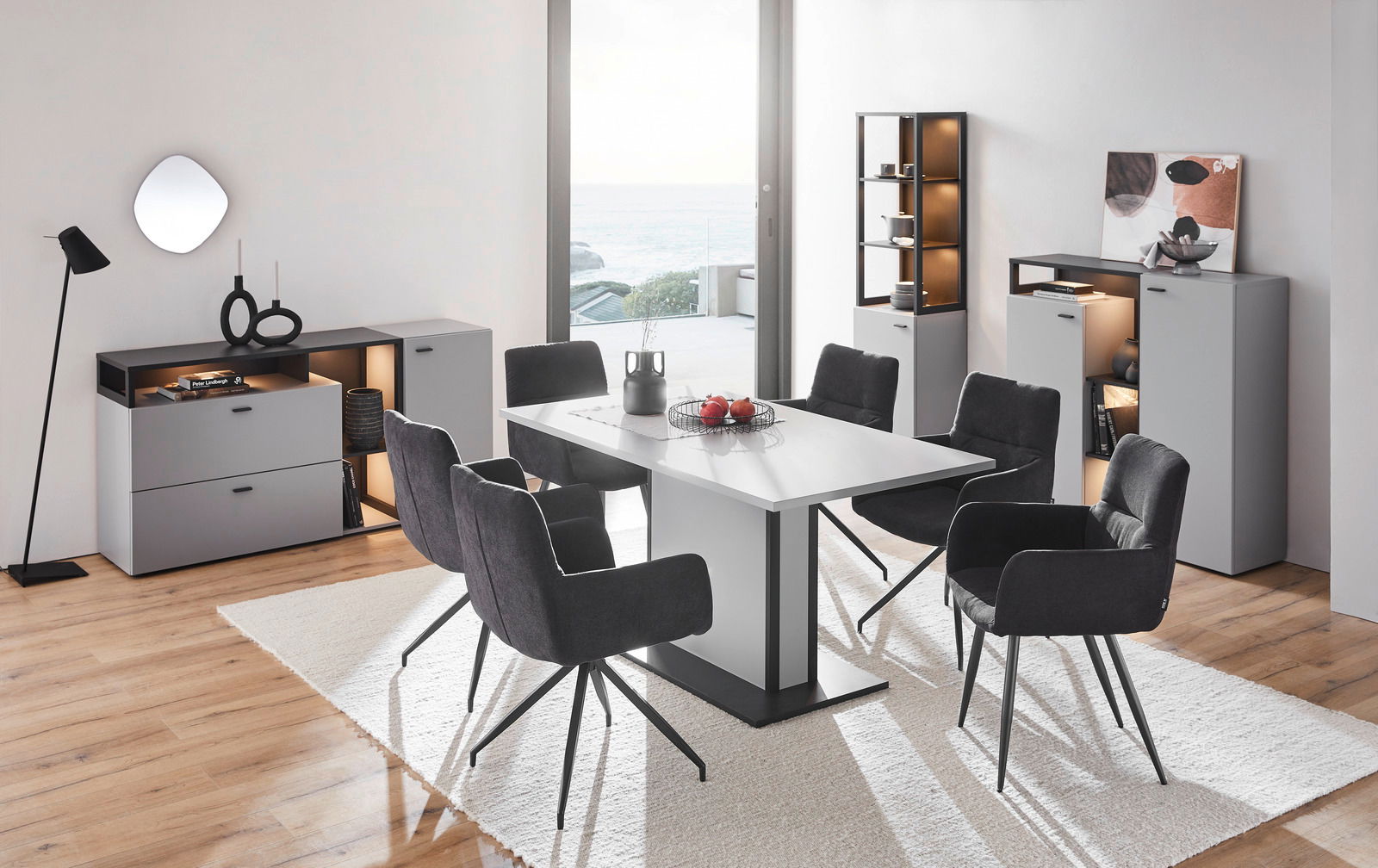 Moderne Esszimmermöbel mit grauen Vitrinen und einem weißen Tisch, umgeben von schwarzen Stühlen, in einem hellen Raum mit Blick auf das Meer. Perspektive von vorne.