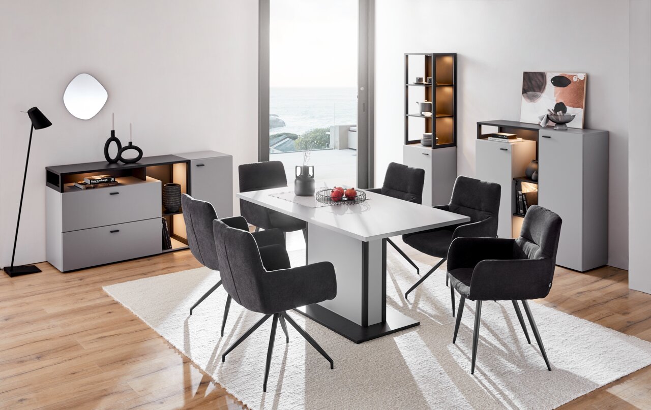 Moderne Esszimmermöbel mit grauen Vitrinen und einem weißen Tisch, umgeben von schwarzen Stühlen, in einem hellen Raum mit Blick auf das Meer. Perspektive von vorne.