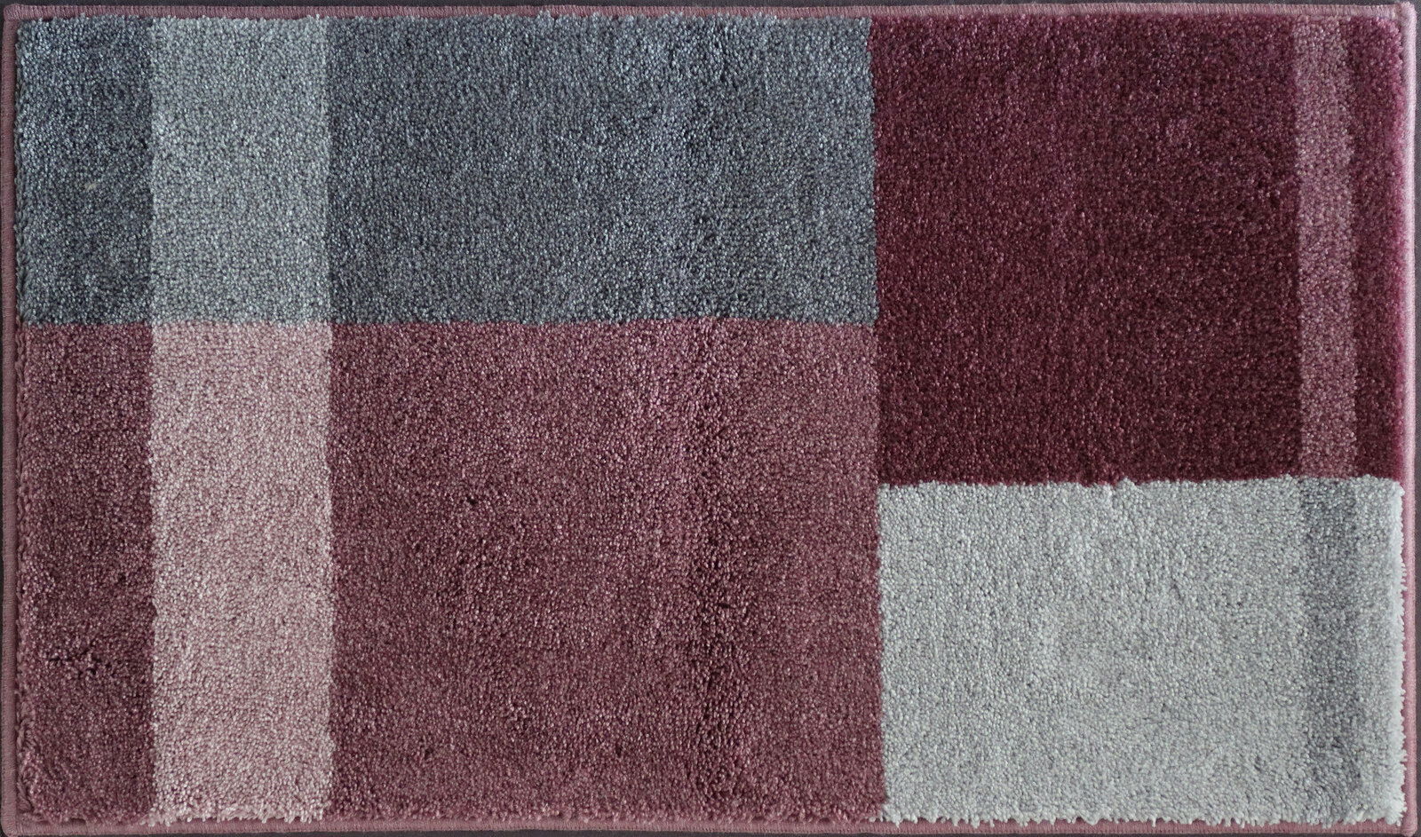 Badteppich in rechteckiger Form mit einem modernen, geometrischen Muster in verschiedenen Grautönen und Rotnuancen, aus der Vogelperspektive fotografiert.