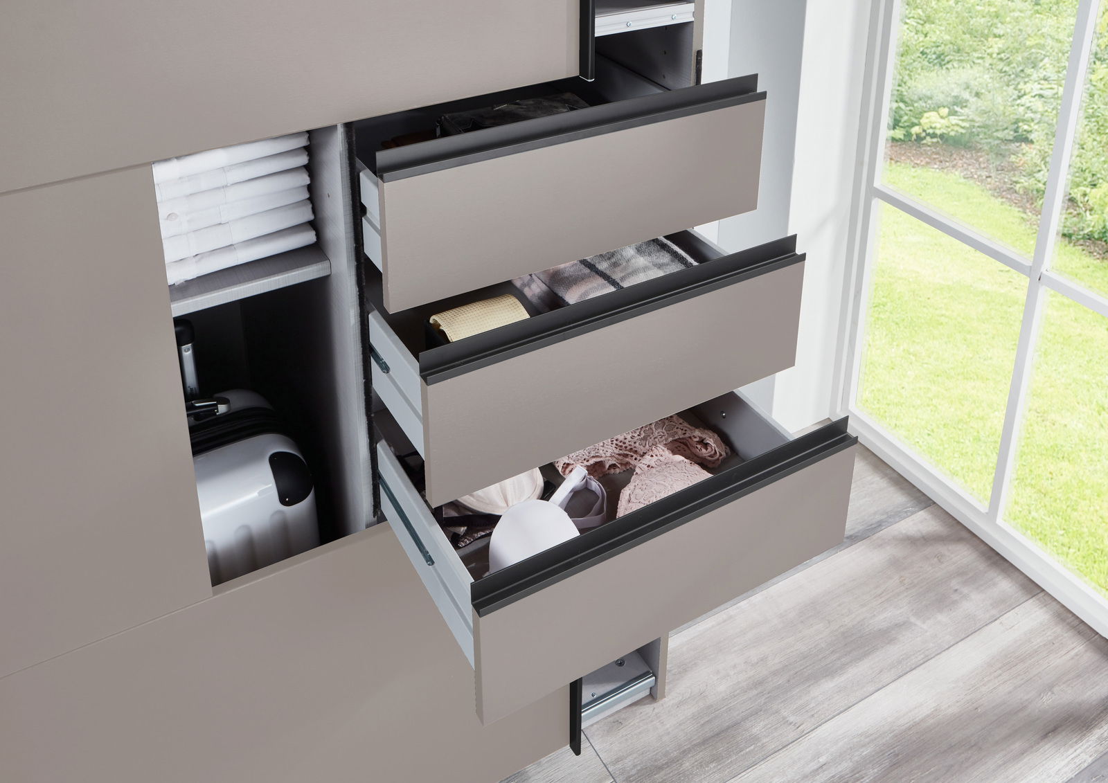 Nahaufnahme eines geöffneten Schwebetürenschranks mit mehreren Schubladen, die Kleidung und Accessoires enthalten. Die Perspektive zeigt den Schrank von der Seite mit Blick auf ein Fenster im Hintergrund.