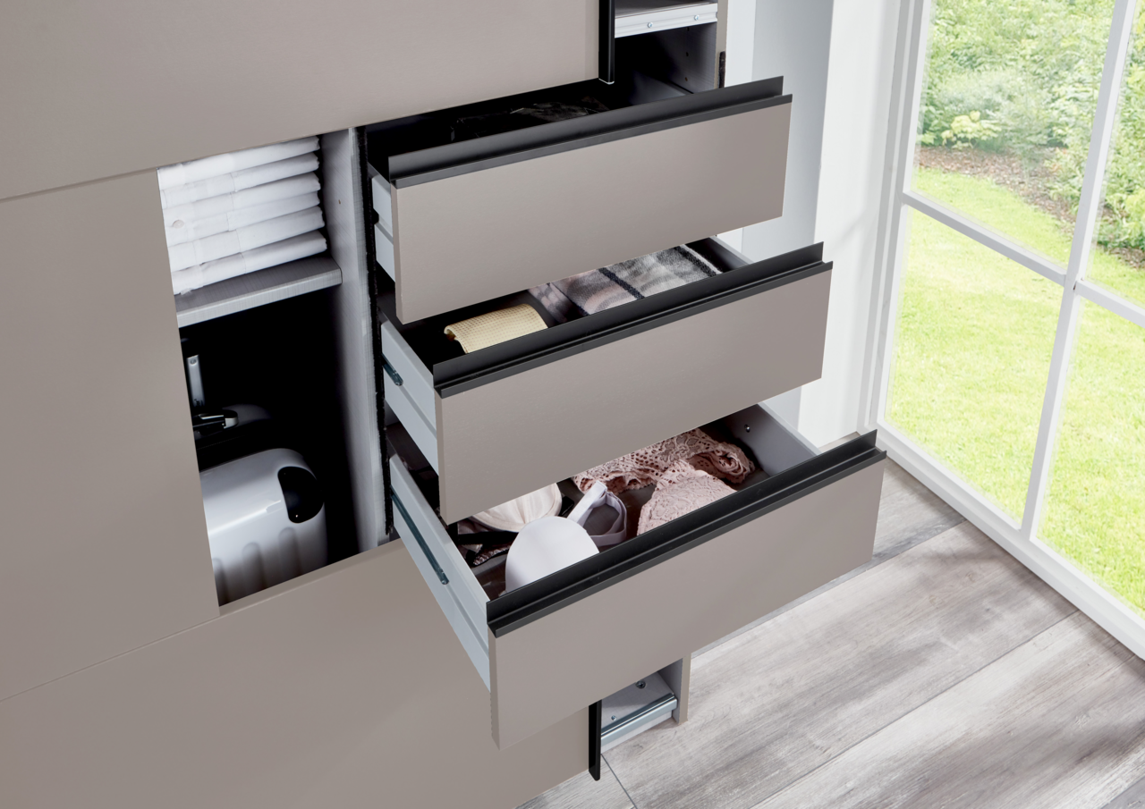 Nahaufnahme eines geöffneten Schwebetürenschranks mit mehreren Schubladen, die Kleidung und Accessoires enthalten. Die Perspektive zeigt den Schrank von der Seite mit Blick auf ein Fenster im Hintergrund.