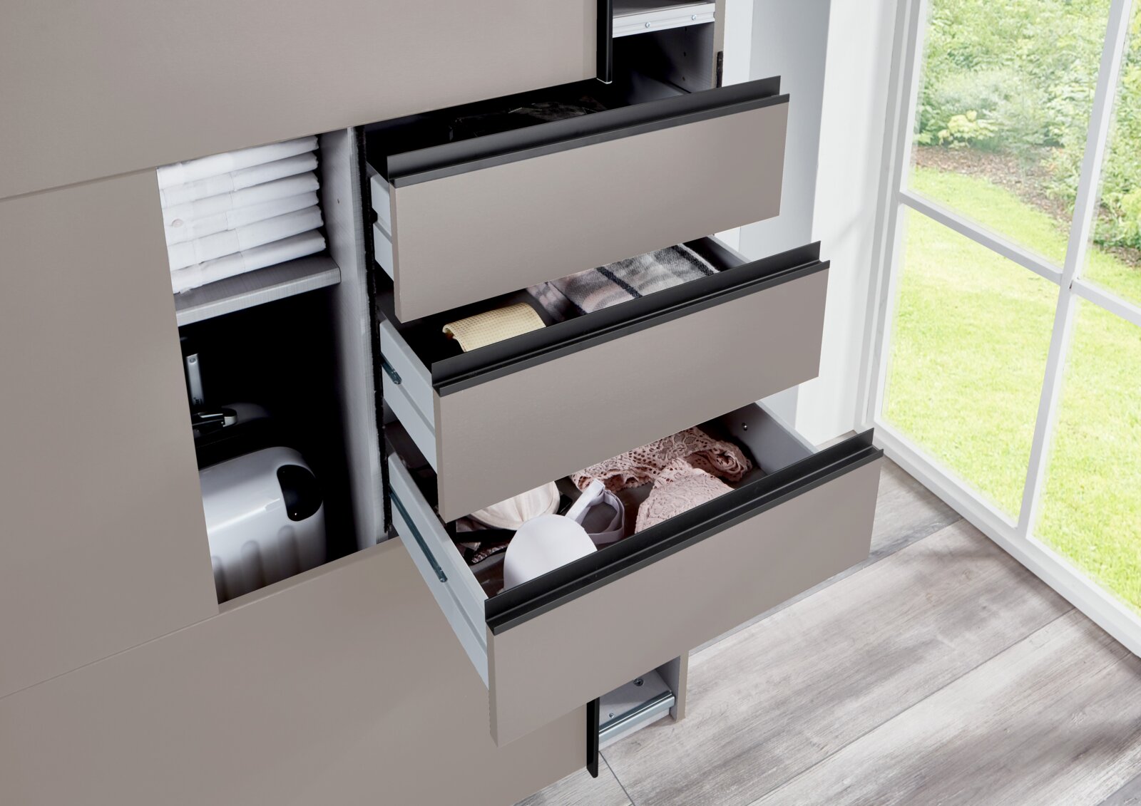 Nahaufnahme eines geöffneten Schwebetürenschranks mit mehreren Schubladen, die Kleidung und Accessoires enthalten. Die Perspektive zeigt den Schrank von der Seite mit Blick auf ein Fenster im Hintergrund.